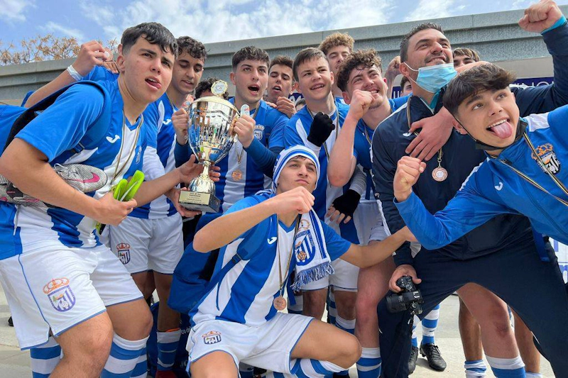A jubilant youth soccer team in blue and white celebrates with a trophy in hand.