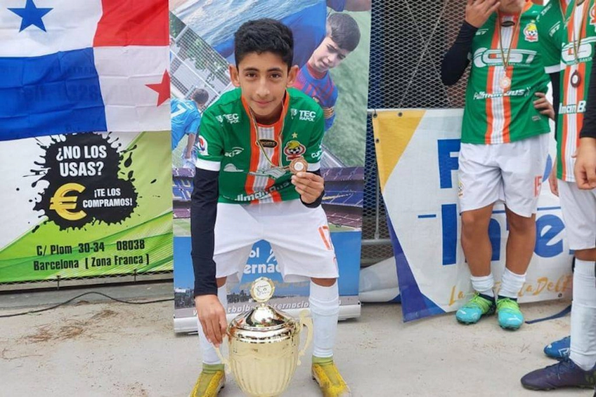 Young soccer player holding a trophy at the Barcelona Youth Cup, flags in the background.