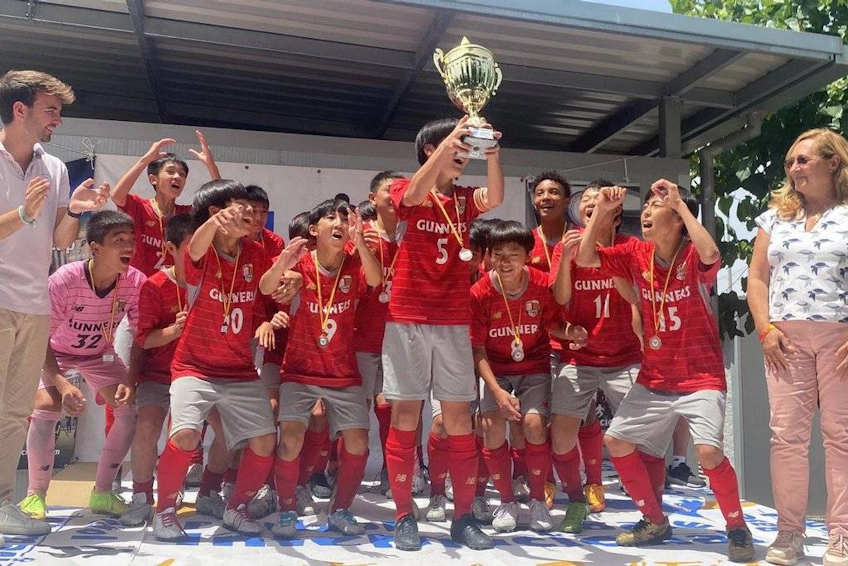 Time de futebol juvenil em êxtase com camisas vermelhas comemorando uma vitória com troféu.