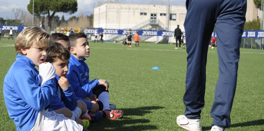 Młodzi piłkarze w niebieskich strojach uważnie słuchają trenera na boisku