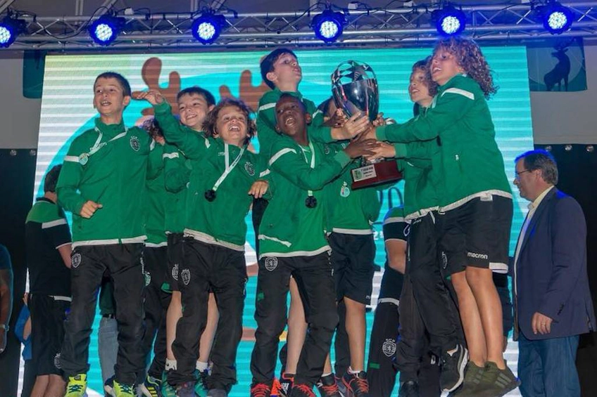 Vrolijk jeugdvoetbalteam in groene jassen tilt een trofee op bij de Miranda Cup zomer voetbaltoernooi.
