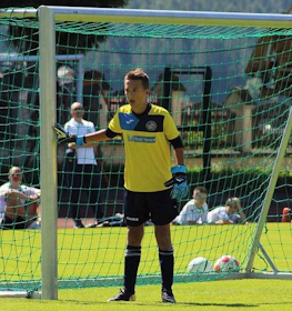 A young goalkeeper in yellow and blue gets ready to defend a goal at a sunny soccer tournament.