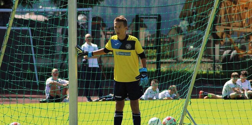 Молодой вратарь в желто-синей форме готовится защитить ворота на солнечном футбольном турнире.