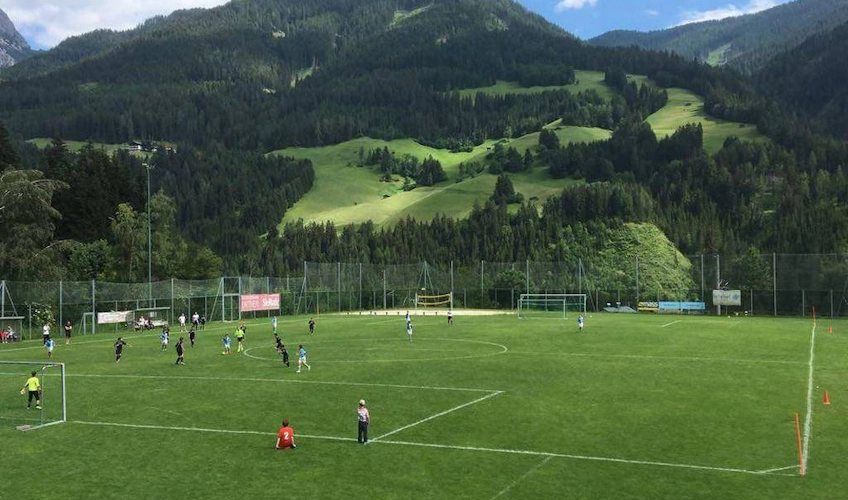 Jalkapallo-ottelu vihreällä kentällä vuoristomaisemalla taustalla
