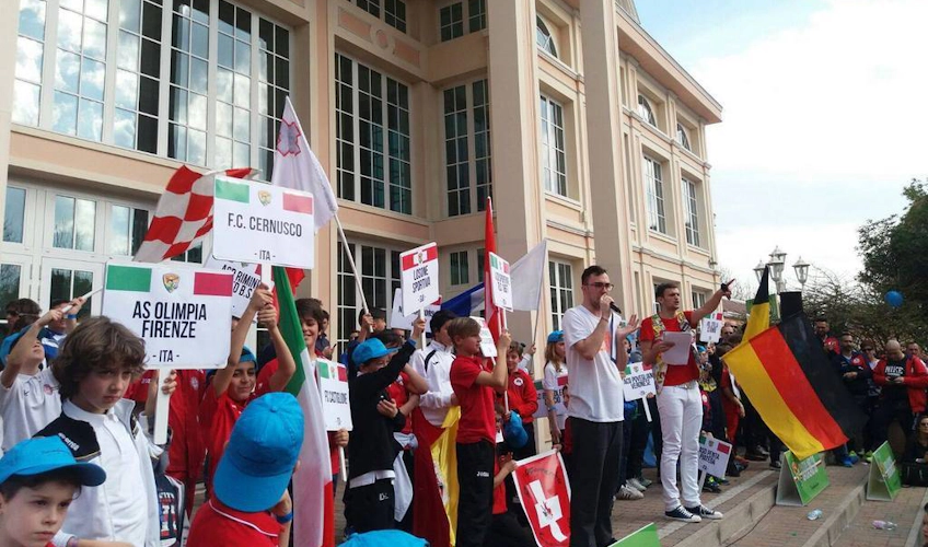 Giovani calciatori entusiasti tengono in alto le bandiere nazionali davanti a un edificio durante la parata della Gardaland Cup.