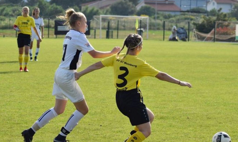 경기 중인 두 명의 여성 축구 선수, 하나는 흰색, 다른 하나는 노란색으로, 푸른 필드에서 공을 두고 경쟁합니다.