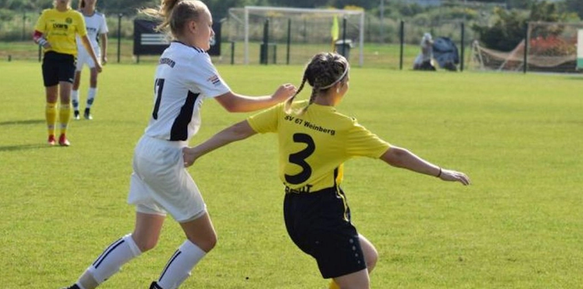 Két női focista egy meccsen, az egyik fehérben, a másik sárgában, küzdenek a labdáért a zöld pályán.