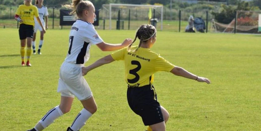两名女足球员在比赛中，一名身穿白色，另一名身穿黄色，正在绿茵场上争夺足球。