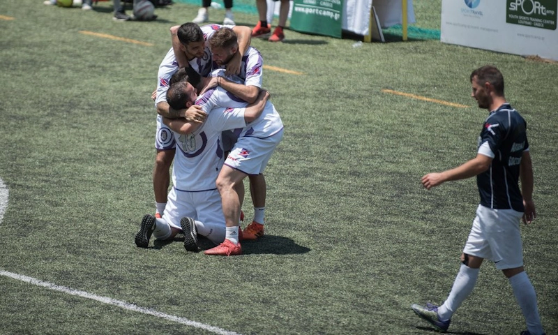 Footballers in white celebrate a goal while a player in black observes, on an artificial pitch.